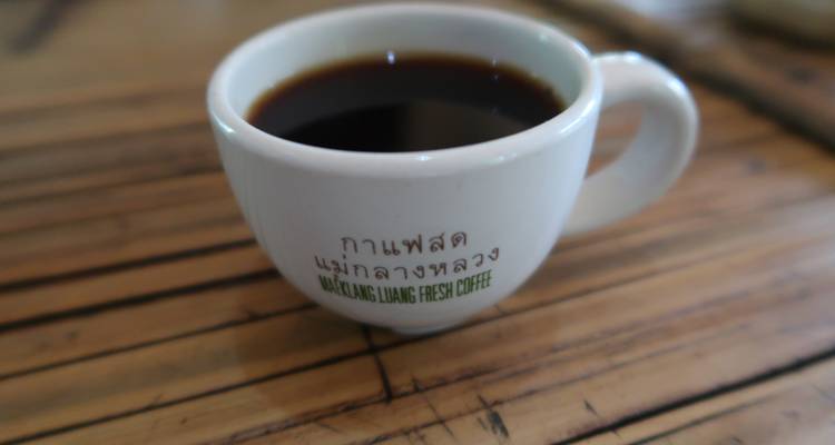Nahaufnahme einer weißen Keramiktasse gefüllt mit schwarzem Kaffee auf einem Bambustisch, Thai-Text auf der Tasse.