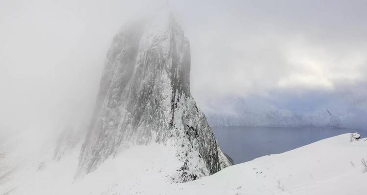 Un pic montagneux spectaculaire couvert de neige émerge d'un brouillard bas au-dessus d'un fjord tranquille.