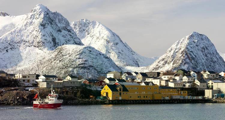 Un bateau de pêche navigue devant un village côtier coloré dominé par des montagnes majestueuses enneigées.