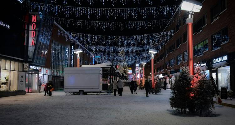 Pedestrian street at night with Christmas lights overhead, small evergreens and shoppers on snowy ground