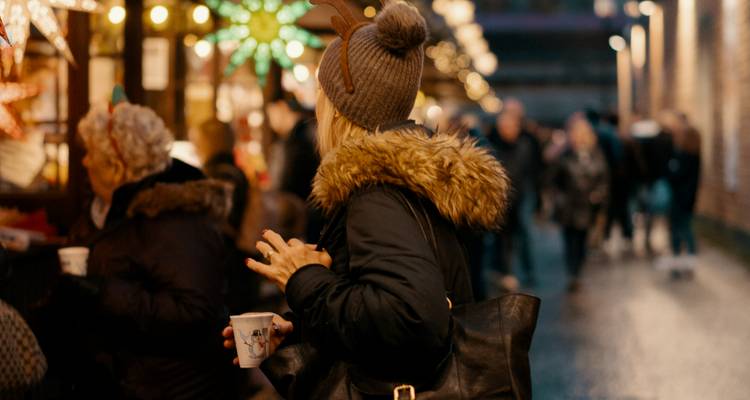 Woman in a fur-lined coat sipping a hot drink at a festive outdoor market crowded with shoppers and warm lights.