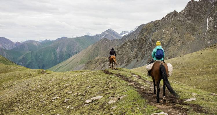 Deux cavaliers à cheval suivant un sentier de crête montagneux avec des pics enneigés devant eux.
