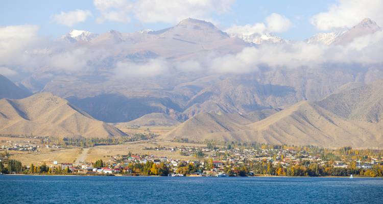 Village lointain sur les rives du Lac Issyk-Kul avec des montagnes imposantes et des nuages épars.