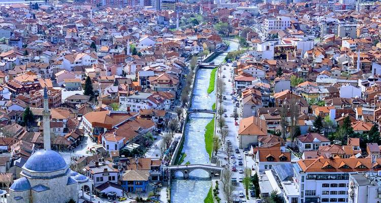 Vue aérienne de la vieille ville de Prizren avec des ponts en pierre traversant une rivière sinueuse bordée de maisons aux toits rouges.