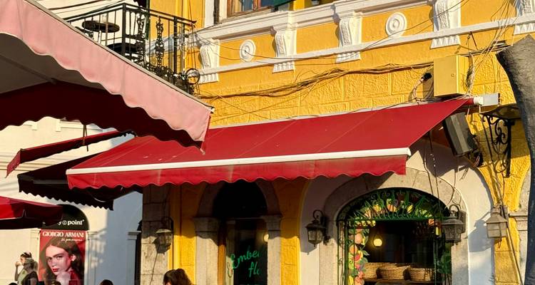 Scène de rue avec une façade d'un jaune éclatant, un auvent rouge et de petites tables de café baignées par la lumière du soleil en fin d'après-midi.