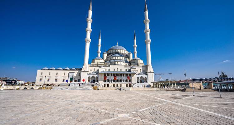 Tagsüber Ansicht der Kocatepe-Moschee, umrahmt von einem großen leeren Vorplatz und leuchtend blauem Himmel.