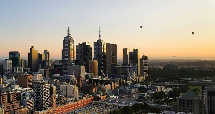 Skyline der Stadt Melbourne bei Sonnenaufgang mit Heißluftballons, die über den Gebäuden schweben, und goldenem Licht am Horizont.