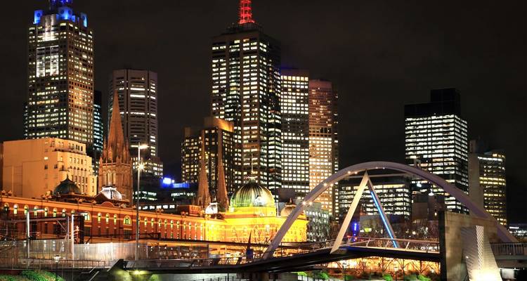 Die beleuchtete Skyline von Melbourne und die Kathedrale bei Nacht, von jenseits des Yarra River mit einer modernen Fussgängerbrücke im Vordergrund betrachtet.