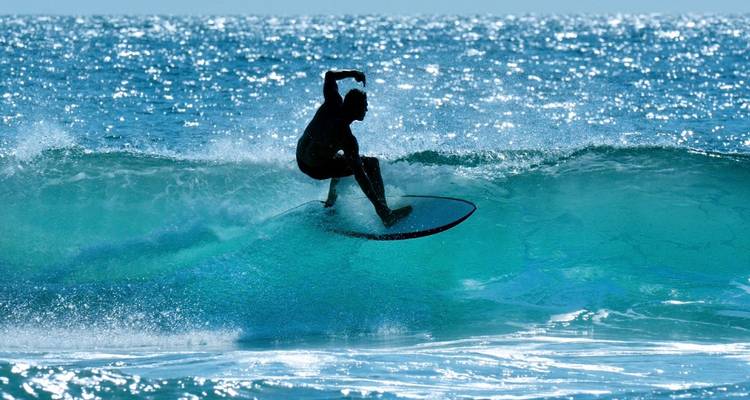 Silhouette eines Surfers, der eine funkelnde türkisfarbene Welle im hellen Sonnenlicht durchschneidet.