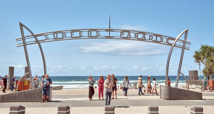 Visitors gather beneath the metallic archway reading 'Surfers Paradise' with ocean waves beyond.