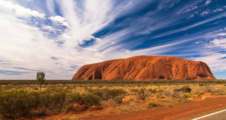 Das ikonische Uluru-Monolith erhebt sich aus flacher Wüstenbuschwelt unter gestreiften Wolken in einem leuchtend blauen Himmel.