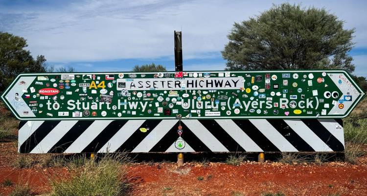 Aufkleber beklebtes Straßenschild, das auf dem roten Schotterweg auf den Stuart Highway und Uluru hinweist.