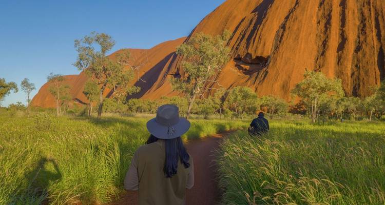Zwei Reisende spazieren bei Sonnenaufgang entlang eines roten Erdwegs neben dem Fuß des Uluru, lange Schatten fallen auf das Gras.