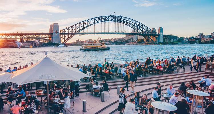 Levendige menigte in de Opera Bar geniet van het uitzicht op de Sydney Harbour Bridge en het Opera House bij zonsondergang.