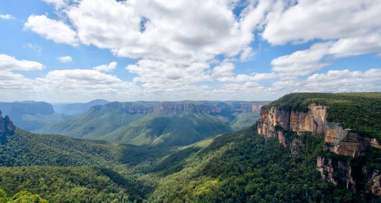 Uitgestrekt uitzicht over beboste valleien en dramatische oranje rotsen van Australië's Blue Mountains onder een lucht vol wolken.