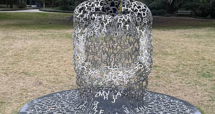Sculpture en lettres métalliques formant une silhouette humaine assise placée dans un parc herbeux.