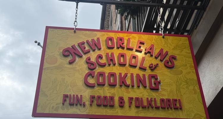 Panneau suspendu de la New Orleans School of Cooking avec une typographie rétro sur un ciel nuageux.