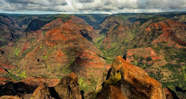 Vue panoramique sur les falaises stratifiées rouges et vertes du Waimea Canyon sous un ciel nuageux spectaculaire.