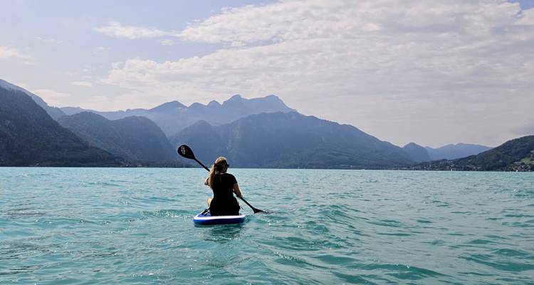 Une femme pratiquant le paddleboard sur un lac alpin turquoise entouré de pics montagneux spectaculaires sous un ciel partiellement nuageux.