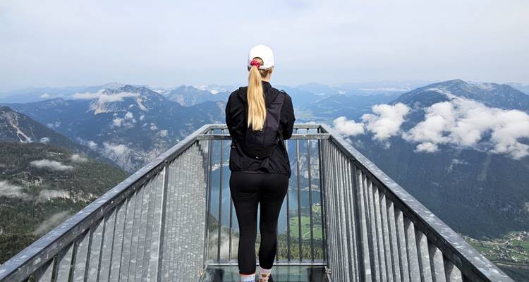 Vue arrière d'une jeune femme se tenant sur une plateforme d'observation panoramique en métal surplombant les nuages et les vallées montagneuses.