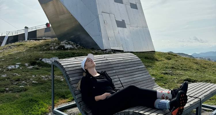 Femme se détendant sur un transat en bois près d'une structure sommitale métallique moderne sur un plateau alpin herbeux.