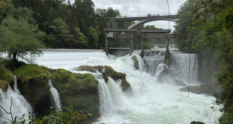 Des cascades puissantes se déversent sur des rochers couverts de mousse en dessous d'un petit barrage et d'un pont en arche dans un ravin forestier.