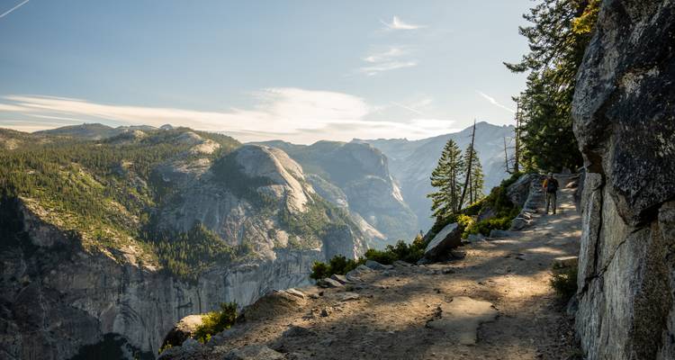 Sentier de haute montagne surplombant des falaises de granit spectaculaires et une vallée boisée sous un ciel dégagé.