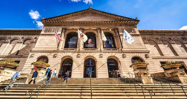 Entrée du grand escalier de l'Art Institute of Chicago avec des visiteurs et des drapeaux agités sous un ciel dégagé.