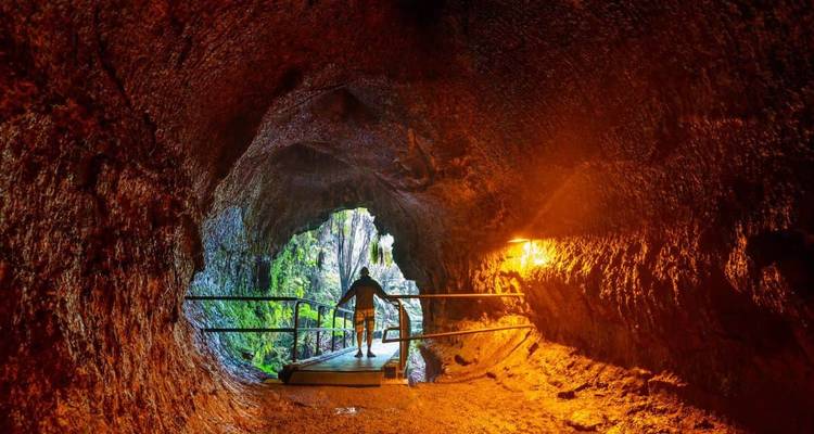 Visitor silhouetted on a boardwalk inside an illuminated lava tube leading to green daylight beyond.