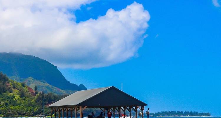 Small pavilion on a pier framed by emerald mountains, blue bay and bright white clouds.