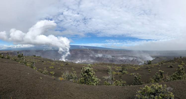Panoramic scene of a steaming volcanic crater with rugged lava fields and patchy vegetation under a bright sky.