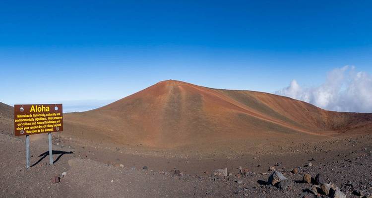 Rust-colored cinder cone rising from a barren volcanic plain with an "Aloha" welcome sign.
