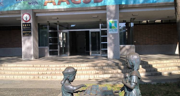 Estatuas de bronce de niños jugando un juego de mesa afuera de la entrada de un edificio moderno iluminado por el sol.