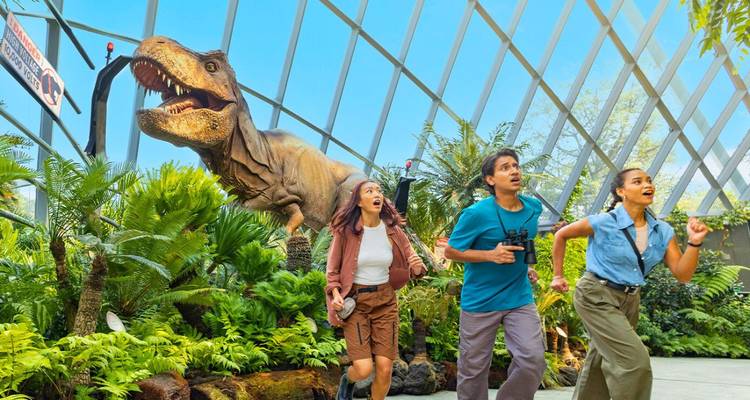 Drei junge Besucher reagieren verspielt auf einen animatronischen T-Rex in einem hellen Glasgewächshaus, das mit tropischen Pflanzen gefüllt ist.