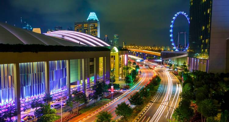 Lebendige Nachtaufnahme der Skyline Singapurs mit Lichtspuren auf einer belebten Straße und dem beleuchteten Singapore Flyer im Hintergrund.