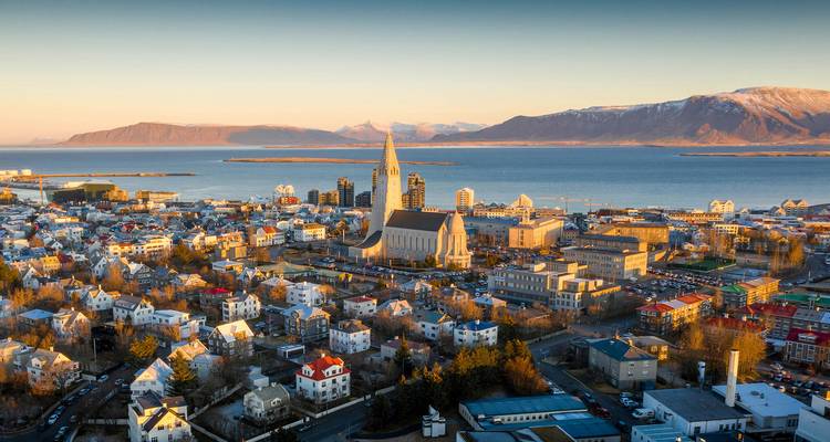 Sonnendurchflutete Luftaufnahme von Reykjavik mit der Hallgrimskirkja im Zentrum, farbenfrohen Dächern und Fjord mit schneebedeckten Gipfeln im Hintergrund