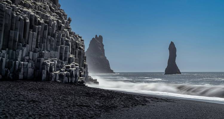 Dramatische Basaltsäulen und schwarzer Sandstrand mit Blick auf Seestapel unter klarem blauen Himmel in Reynisfjara
