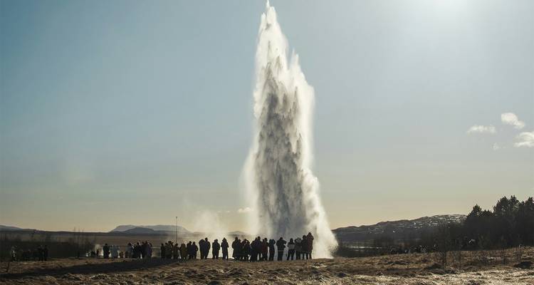 Zuschauer beobachten einen imposanten Geysir, der Wasser und Dampf bei sonnigem Wetter hoch in den Himmel schießt.