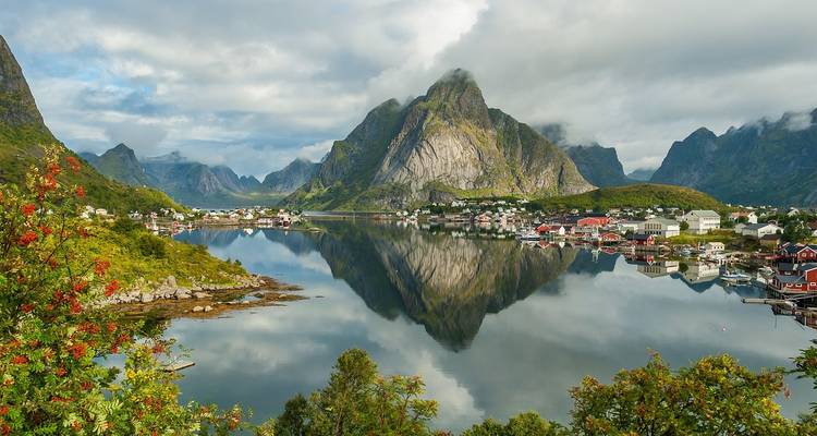 Fiordo espejo tranquilo reflejando montañas espectaculares y un pequeño pueblo pesquero bajo un cielo parcialmente nublado