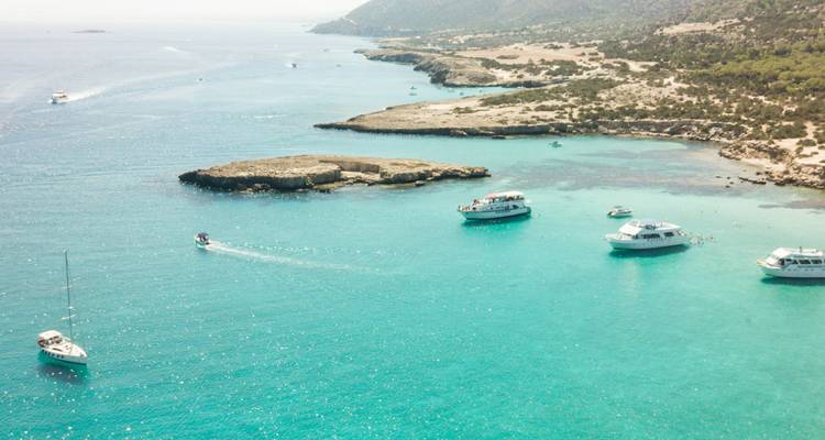 Vista aérea de una bahía turquesa brillante salpicada de barcos de recreo frente a una península chipriota escarpada y boscosa
