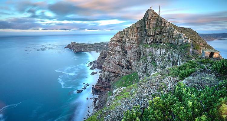 Steep sheer cliff of Cape Point crowned by a lighthouse, dropping to swirling blue ocean at dawn.
