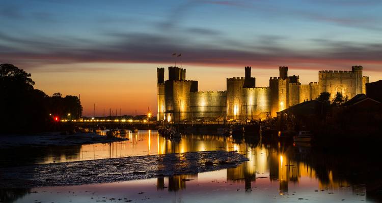 El Castillo de Caernarfon iluminado al atardecer, reflejado en aguas tranquilas de la marea bajo un cielo de puesta de sol vivida.