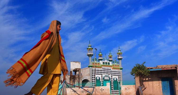 Un homme vêtu d'un châle traditionnel marche devant une petite mosquée rurale aux motifs colorés sous un ciel bleu éclatant parsemé de nuages légers.