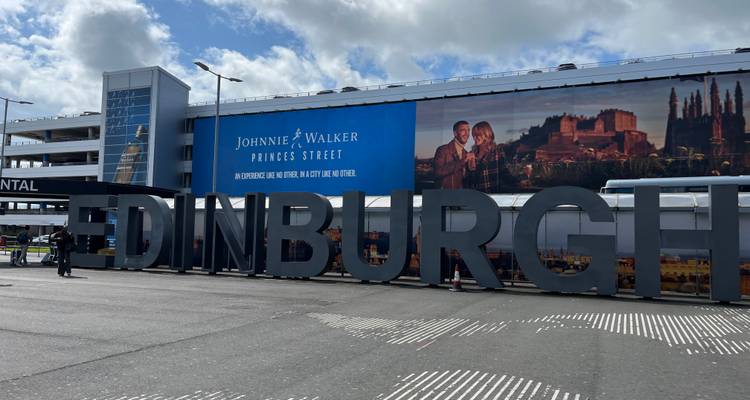 Grand panneau EDINBURGH à l'extérieur du terminal de l'aéroport avec voyageurs et décor publicitaire.