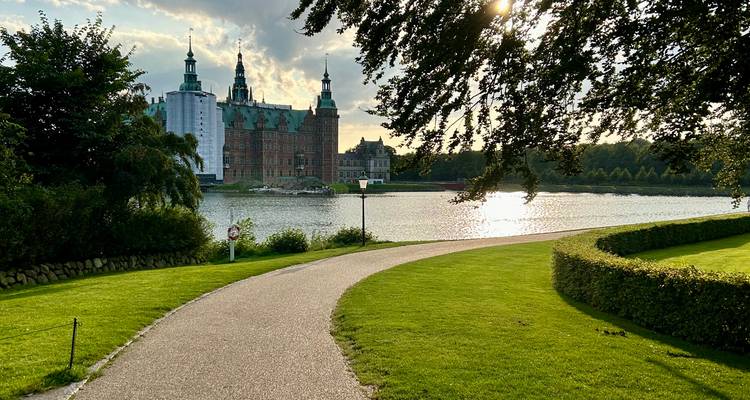 Kronkelend pad door verzorgd parklandschap leidt naar kasteeltje aan het meer, verlicht door het licht van de late middag.