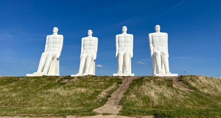 Vier imposante witte 'Mannen aan Zee' standbeelden zitten op een graszige duin onder een helderblauwe hemel.