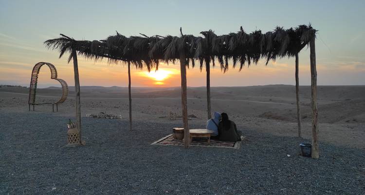 Un couple est assis sous un dais de frondes de palmier et regarde le soleil se coucher sur les collines désertiques ondulantes.