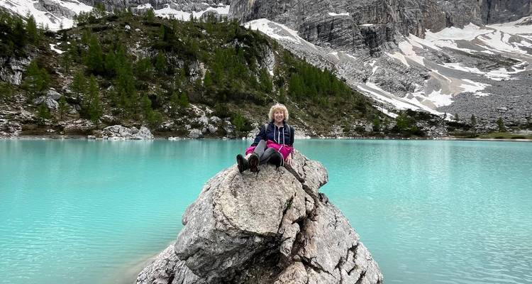 Ein Reisender sitzt auf einem Felsbrocken und überblickt einen beeindruckenden türkisfarbenen Alpensee, der von felsigen Dolomitengipfeln umgeben ist.