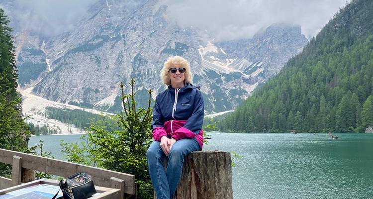 Ein sitzender Reisender genießt den ruhigen Blick auf einen smaragdgrünen Alpensee, der von bewaldeten Bergen an einem bewölkten Tag umgeben ist.