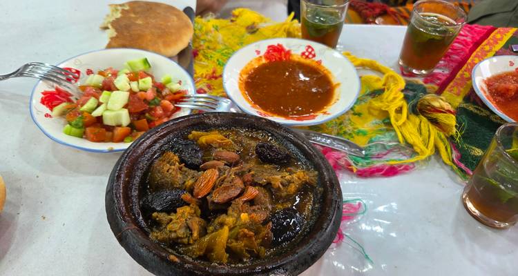 Traditionele Marokkaanse tajine met pruimen en amandelen naast verse salade, brood en thee op een kleurrijke tafel.